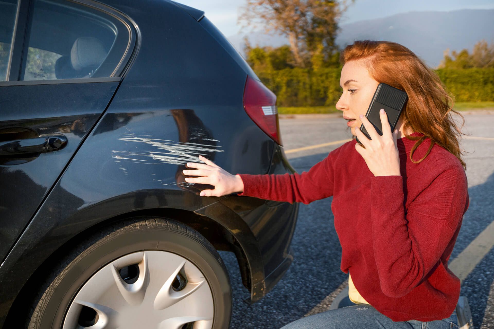 Eine Frau in einem roten Pullover kniet neben einem schwarzen Auto und deutet auf einen auffälligen Kratzer, während sie telefoniert.