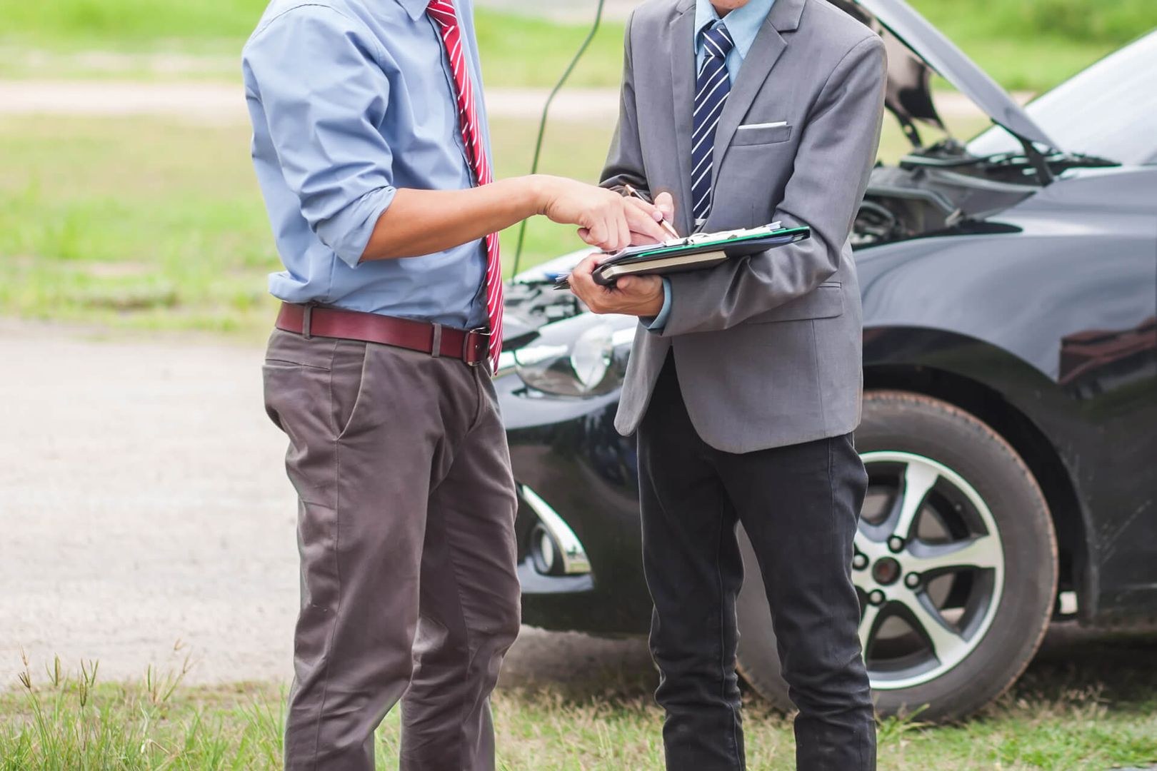 Zwei Männer diskutieren über Autoreparaturen, einer in blauem Hemd und roter Krawatte, der andere im Anzug mit einem Klemmbrett in der Hand, neben einem schwarzen Auto.