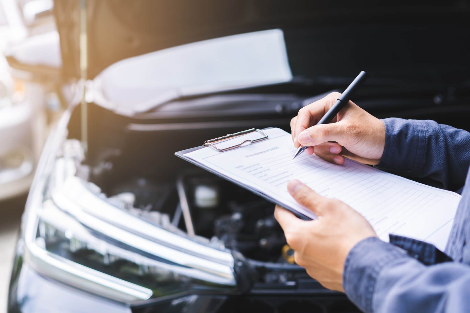 Ein Mechaniker hält ein Klemmbrett und einen Stift in der Hand, während er einen Automotor bei geöffneter Motorhaube inspiziert und die Ergebnisse für die Wartung dokumentiert.