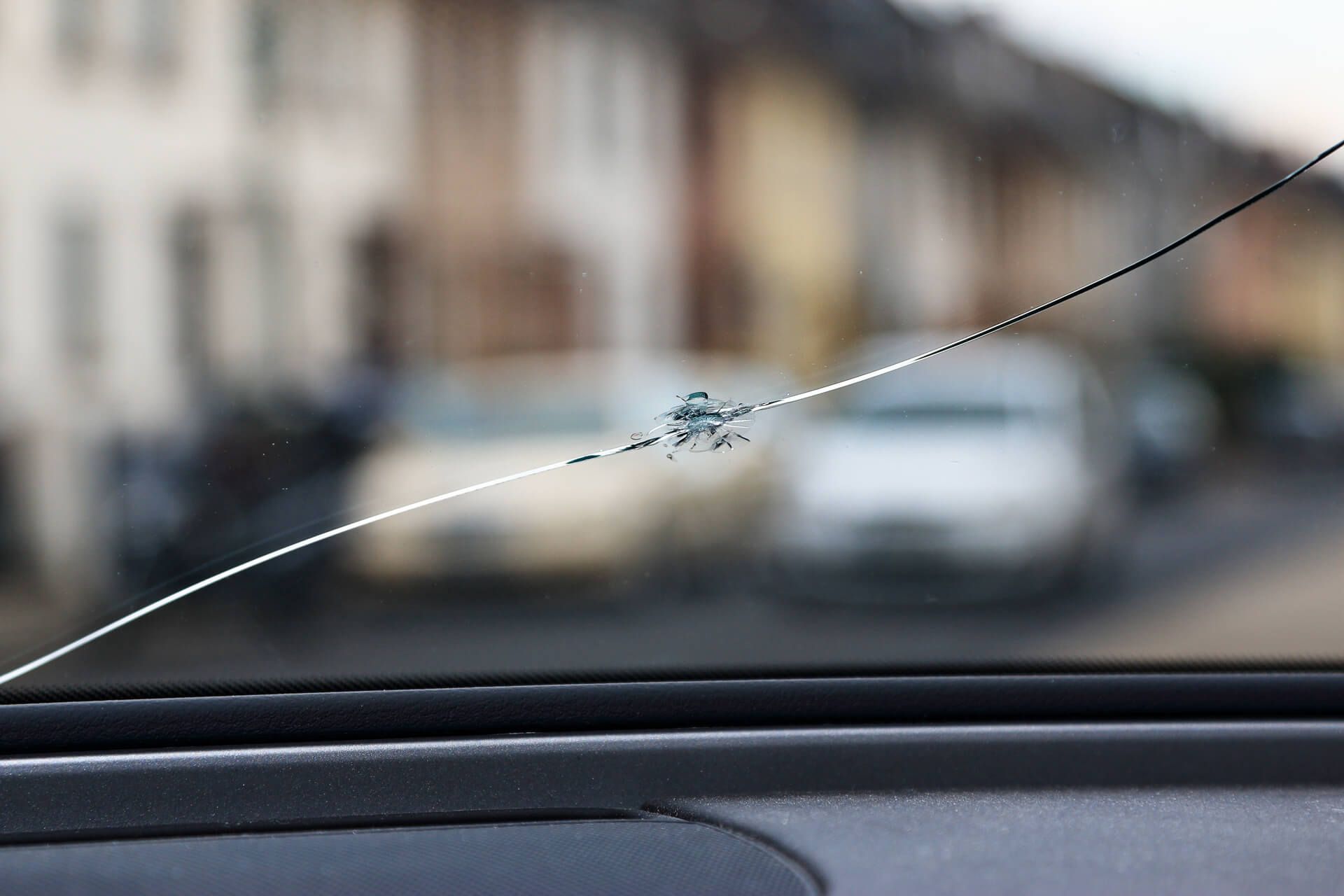 Nahaufnahme einer gesprungenen Autoscheibe mit einem deutlichen Chip; im Hintergrund sind eine unscharfe Wohnstraße und geparkte Autos zu sehen.